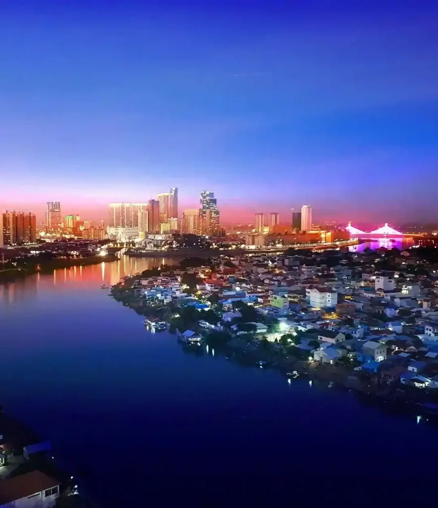 Aerial view of the Phnom Penh skyline in Cambodia at blue hour, showing a curving river (likely the Mekong or Tonle Sap) in the foreground and a cluster of modern, illuminated skyscrapers reflecting off the water.